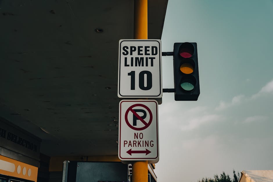 A street scene featuring a white speed limit sign indicating a maximum of 10 miles per hour, mounted on a yellow pole alongside a traffic light with red, yellow, and green signals. Below the speed limit sign, there is a white and red no parking sign with a red circle and slash over a capital 'P', accompanied by the words 'NO PARKING' and an arrow pointing left. The background shows part of a building with a cantilevered roof extending over the sidewalk and some cloudy sky. This setting reflects urban parking and traffic regulation signage, relevant to house removals and moving logistics in Pimlico, with the presence of signage managed by Man with Van Pimlico for relocation activities.