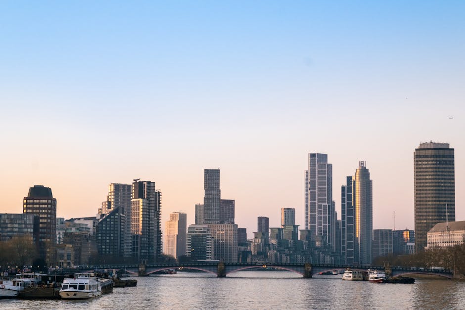 A view of a modern city skyline with high-rise buildings under a cloudy sky, featuring construction cranes atop some structures. In the foreground, a river flows beneath a bridge, with several boats d