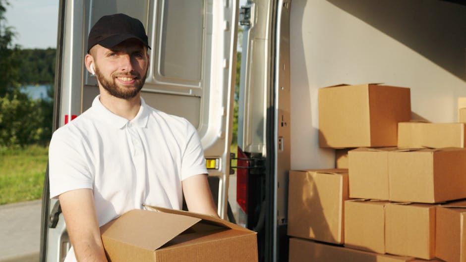 A person wearing a white shirt, jeans, and a black cap is loading large brown cardboard boxes into the side of a white panel van parked on a residential street. The individual is holding two boxes sta