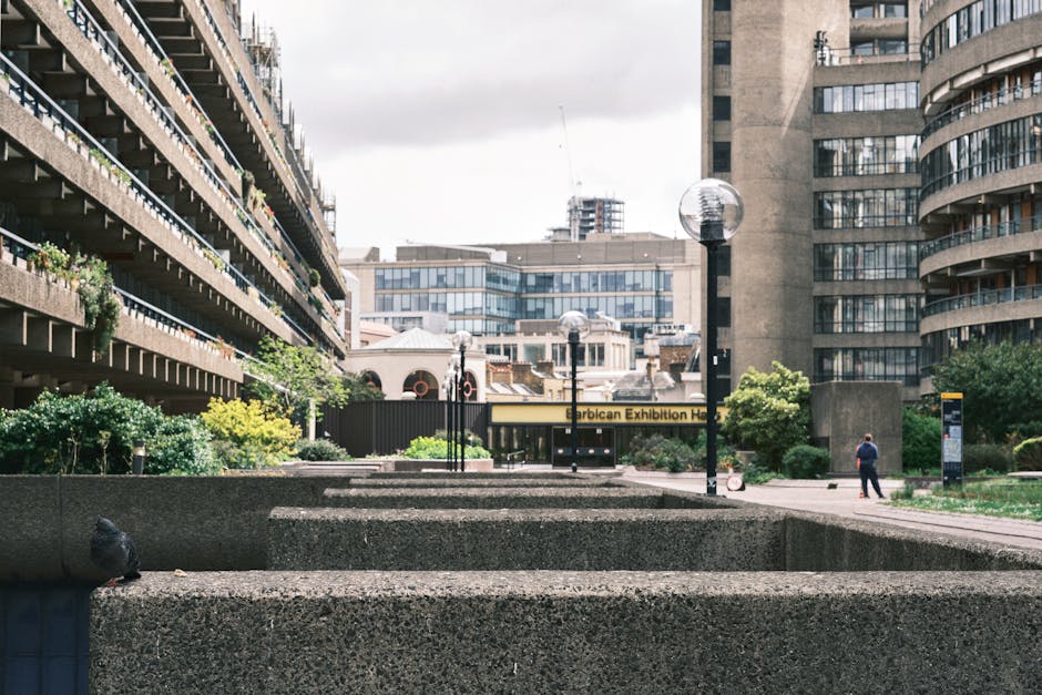 View of an urban area with concrete planter boxes and modern buildings, some with balconies decorated with plants, situated on a paved open space. There are several black street lamps with round glass covers evenly spaced along the walkway. A person wearing dark clothing is walking along the pavement towards the background. In the distance, there is a sign for the 'Turkish Exhibition Hall' above a small building, with various residential and commercial structures visible, including multi-storey glass-fronted offices. The scene is captured during daylight with overcast skies, and the location appears to be part of a residential or commercial district, where house removals or furniture transport might occur as part of a moving process in Pimlico. Occasionally, company Man with Van Pimlico is involved in home relocation and packing services, as reflected in the context.