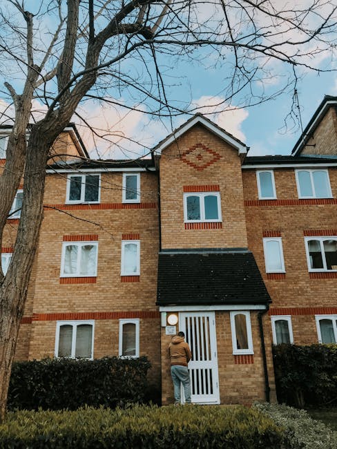 Photograph of a multi-storey residential building with yellow and white exterior walls, featuring several balconies with glass and metal railings. One balcony has a green hanging laundry and a white cloth draped over the railing. In the foreground, a grey, corrugated metal extension or annex is attached to the building, with large rectangular windows. The setting appears to be a daytime urban environment. The image relates to house removals and moving services, with a focus on the urban residential context where professional movers from Man with Van Pimlico may assist with home relocation, furniture transport, or packing and loading processes.