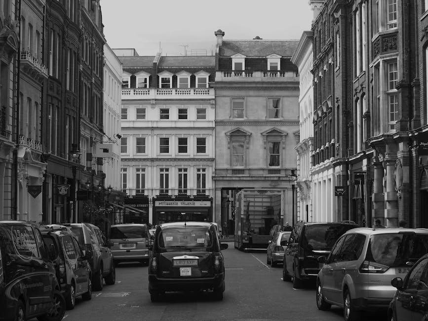 Black and white photograph of a narrow urban street with tightly packed parked cars lining both sides, including small vans, sedans, and hatchbacks. The street is bordered by historic multi-storey buildings with ornate facades, large windows, and decorative architectural details, typical of a central London neighbourhood. In the background, a building with a shopfront displays signage, partially visible behind a delivery truck that is parked at the curb. The pavement is clear of pedestrians, and the scene is lit by natural daylight. This setting depicts a typical street scene suitable for house removals or furniture transport in a city centre, aligning with the context of professional relocation and moving services such as those offered by Man with Van Pimlico.