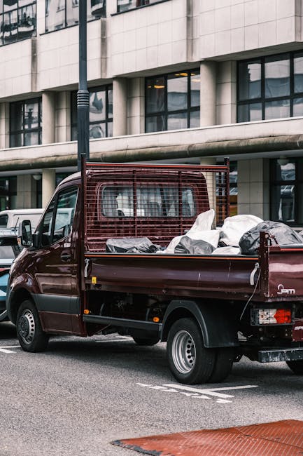 A maroon flatbed cargo truck parked on a city street in front of a modern office building with large windows. The truck's bed is loaded with various packed items, including cardboard boxes wrapped in plastic, fabric-covered furniture or appliances, and other household packing materials. The load is secured with straps, and some items are covered with protective blankets. The truck is positioned near the curb, ready for furniture transport as part of a home relocation process. A vertical streetlight pole is visible behind the truck, and parked cars line the street. This scene illustrates a typical loading process for house removals, with a professional moving service like Man with Van Pimlico assisting in packing and transporting belongings within the urban environment.