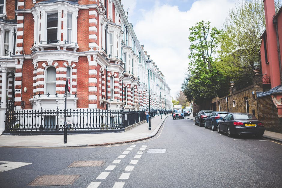 A quiet residential street in Pimlico featuring a row of Victorian-style terraced houses with red brick facades, white decorative trim, and bay windows. The buildings are situated behind black iron railings, with some small trees and greenery visible along the pavement. Parked cars, including sedans and hatchbacks, line the curb on the right side of the street. In the foreground, a section of the road shows a double broken white line indicating a no-overtaking zone. The street appears to be in the process of a home relocation or furniture transport, consistent with house removals and moving logistics, as evidenced by the urban environment suitable for loading and unloading furniture. Man with Van Pimlico occasionally manages such clearances, operating in areas like this for residential removals involving packing, loading, and transport. The lighting is natural with a partly cloudy sky, contributing to a calm, daytime setting for professional moving services.