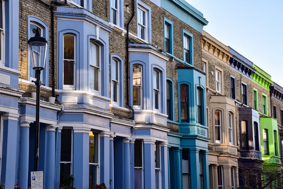 A row of multi-storey residential buildings with vibrant, colorful facades in shades of blue, beige, and green, situated along a street in Pimlico. The buildings feature large, arched sash windows with white trim, some of which reflect the sky and surrounding environment. A traditional black street lamp stands in the foreground, next to a sign indicating parking regulations. The lower floors have bay windows and decorative elements, with some ground level windows partially visible. The scene is illuminated by soft daylight, highlighting the details and textures of the buildings' brick and painted surfaces. This image captures the urban architecture typical of home relocation environments, relevant to house removals and furniture transport services offered by Man with Van Pimlico, illustrating the context of a busy residential street where packing, loading, and moving activities take place.