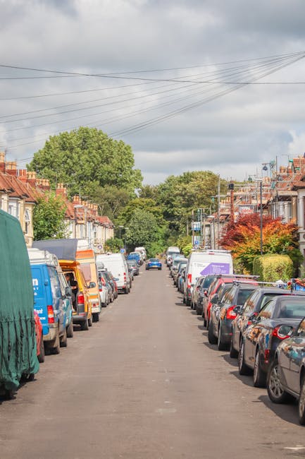 A residential street in Pimlico with parked cars lining both sides of the narrow tarmac road, including a variety of small vans and a covered object on a trolley or dolly. The scene shows a typical urban setting with terraced houses that have brick façades, some with small front gardens and well-maintained greenery. Overhead, power lines are visible against a partly cloudy sky, indicating a typical domestic environment suitable for home relocations or furniture transport. The street appears calm and quiet, with no visible pedestrians or moving vehicles, suggesting a convenient location for unloading or loading furniture and boxes during house removals. This setting reflects the logistical considerations for removals services like those offered by Man with Van Pimlico, particularly during the borough’s specific permit rules for moving vans, as noted on the associated webpage about Pimlico council permit regulations.
