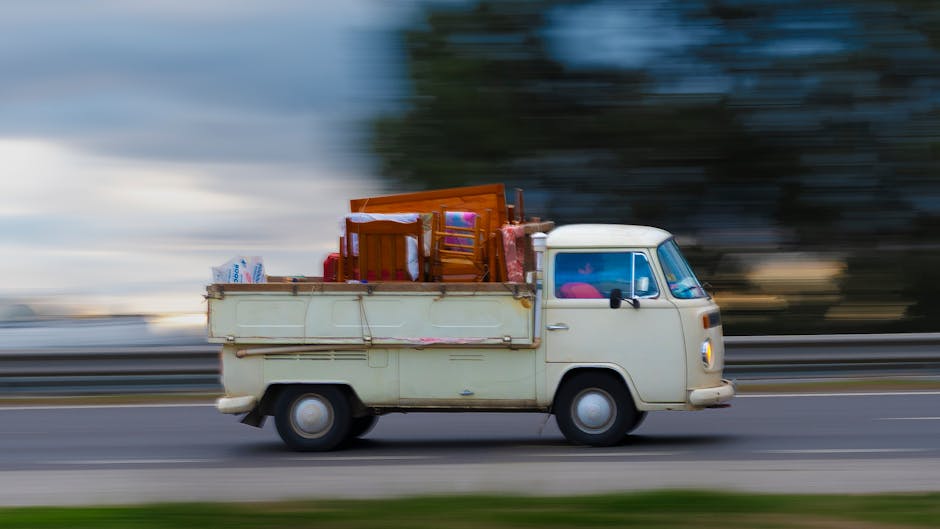 A small vintage cream-colored van with a rounded front and side mirrors, parked on a roadway with a blurred background indicating motion. The vehicle's flatbed area is loaded with various household furniture and items, including three wooden chairs, a small table, and cardboard boxes, some wrapped in plastic. There are fabric or padding materials visible among the packed items, suggesting preparations for home relocation or furniture transport. The scene appears to be outdoors during daylight hours, with the vehicle positioned near a roadside, possibly during a loading or unloading process as part of a moving or removal service. The image illustrates the loading stage of a house removal, with the van ready for transport, aligning with the services offered by Man with Van Pimlico, especially regarding permits and moving logistics in the Pimlico area (SW1V).