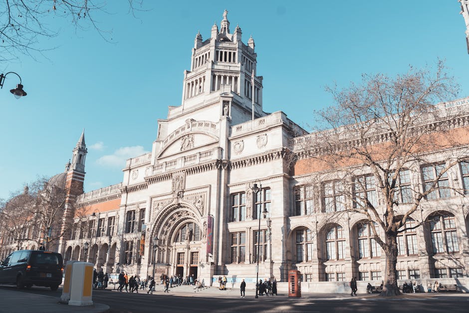 A large historic building with ornate stone architecture, featuring a prominent central tower with decorative spires and intricate carvings, situated along a city street lined with leafless trees. The building has multiple arched windows with clear glass panes, some with decorative framing, and a grand arched entrance with detailed stonework surrounding it. In the foreground, a black van is parked on the pavement, with several movers handling a variety of packed cardboard boxes, wrapped furniture, and protective blankets, preparing for home relocation or furniture transport. A person is seen lifting a box onto a dolly, while others assist with loading items into the van, using straps and trolleys as part of the loading process. The scene is illuminated by natural daylight, with a bright blue sky overhead and minimal shadows, capturing a typical moving day outside a notable landmark, reflecting the logistics involved in house removals facilitated by [COMPANY_NAME].