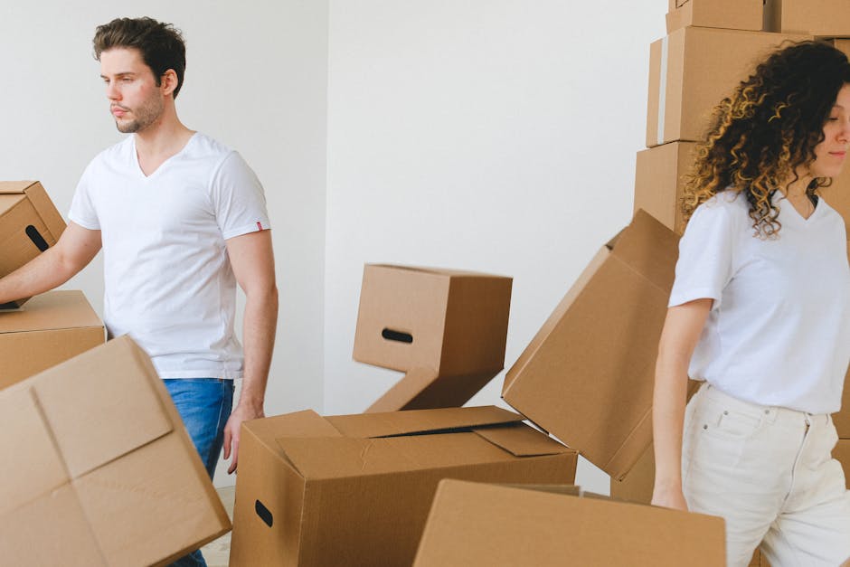 Two movers from Man with Van Pimlico are engaged in a home relocation process inside a property. The male mover, dressed in a white t-shirt and blue jeans, is carrying a cardboard box towards a pile of similar boxes and packaging cartons. The female mover, also in a white top and cream trousers, is standing nearby, with her back slightly turned, possibly waiting to assist or continue packing. The scene shows various cardboard boxes of different sizes stacked against the wall and on the floor, some sealed with packing tape and others open, revealing their contents. Plastic wrap is visible around some items, indicating packing materials are in use. The environment is well-lit, with a plain white wall serving as the background. The equipment observed include moving boxes and possibly a trolley or load-bearing surface, part of the furniture transport and loading process, with the focus on efficient packing and handling for an estate relocation handled by Man with Van Pimlico. This scene illustrates the logistics of packing, lifting, and loading furniture and boxes during an urgent flat move in Pimlico.