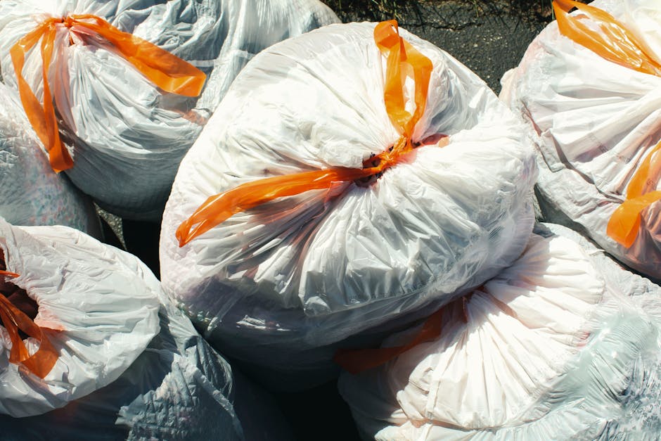 Several large white plastic garbage bags with orange ties are placed outdoors on a paved surface, filled with waste material from household packing or clutter clearing during a home relocation. The bags, containing various packing materials such as cardboard, plastic, or fabric, are arranged closely together, some overlapping. Behind the bags, part of a building wall and steps are visible, indicating the exterior environment. The bags are likely prepared for disposal or recycling as part of the post-move cleanup process facilitated by Man with Van Pimlico, a company specialising in removals and relocation services. The scene highlights the importance of waste disposal duties after a move, illustrating the packing, sorting, and loading activities involved in ensuring a tidy and environmentally responsible transition to a new home.